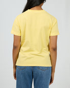 A person shown from the back wearing a pale yellow short-sleeve crewneck T-shirt and blue jeans against a light gray studio background.