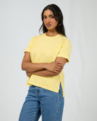 A woman modeling a pale yellow short-sleeve t-shirt with side slits, paired with blue jeans, against a light gray studio background.