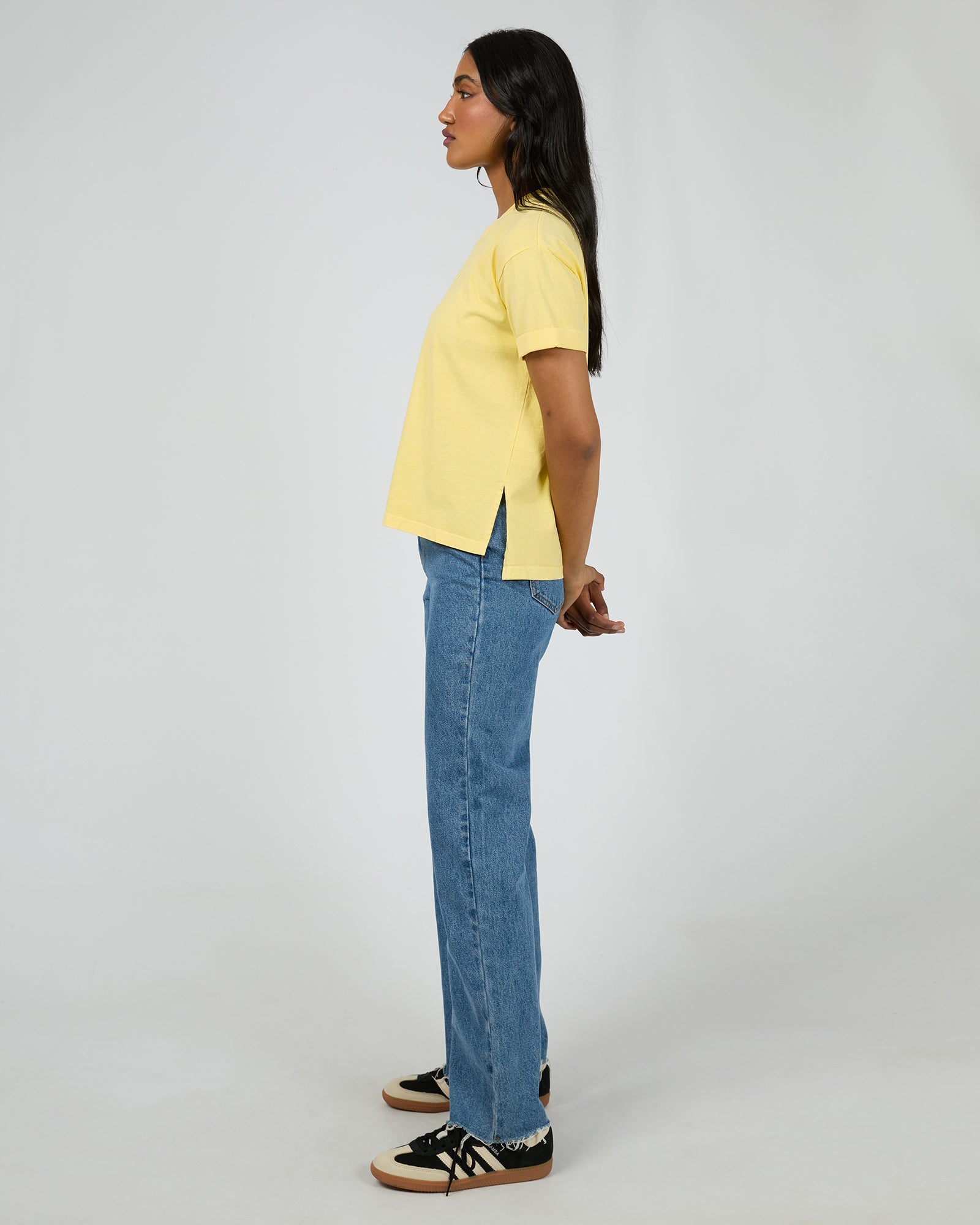 A woman shown in profile wearing a pale yellow short-sleeve top, blue jeans, and black sneakers against a plain light gray studio background.