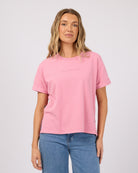 A woman wearing a light pink short-sleeve t-shirt with 'ALL ABOUT ME.' printed in small dark letters on the chest, paired with blue jeans, against a light gray studio background.