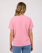 A woman viewed from behind wearing a light pink short-sleeve t-shirt and blue jeans against a plain light gray background.