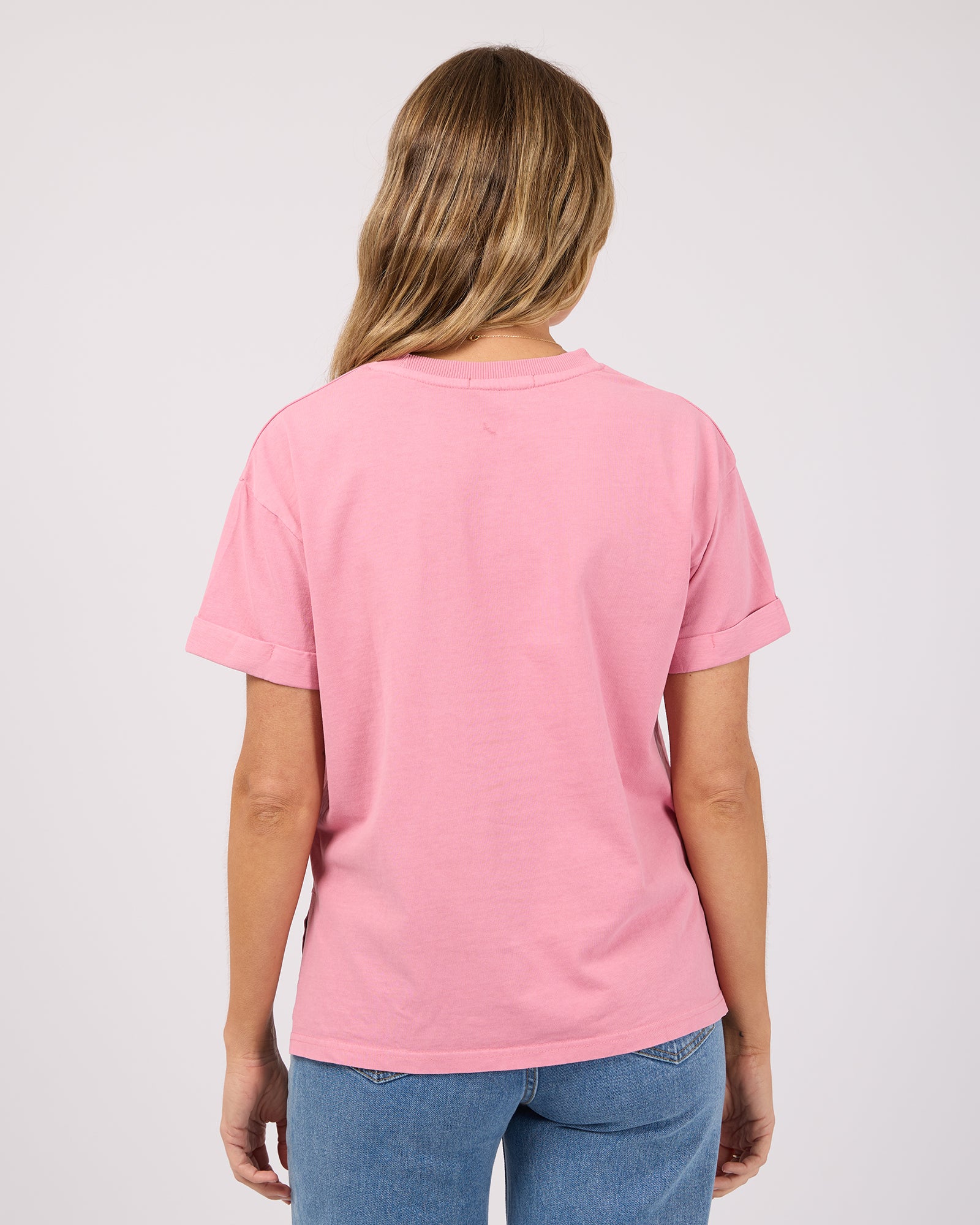 A woman viewed from behind wearing a light pink short-sleeve t-shirt and blue jeans against a plain light gray background.