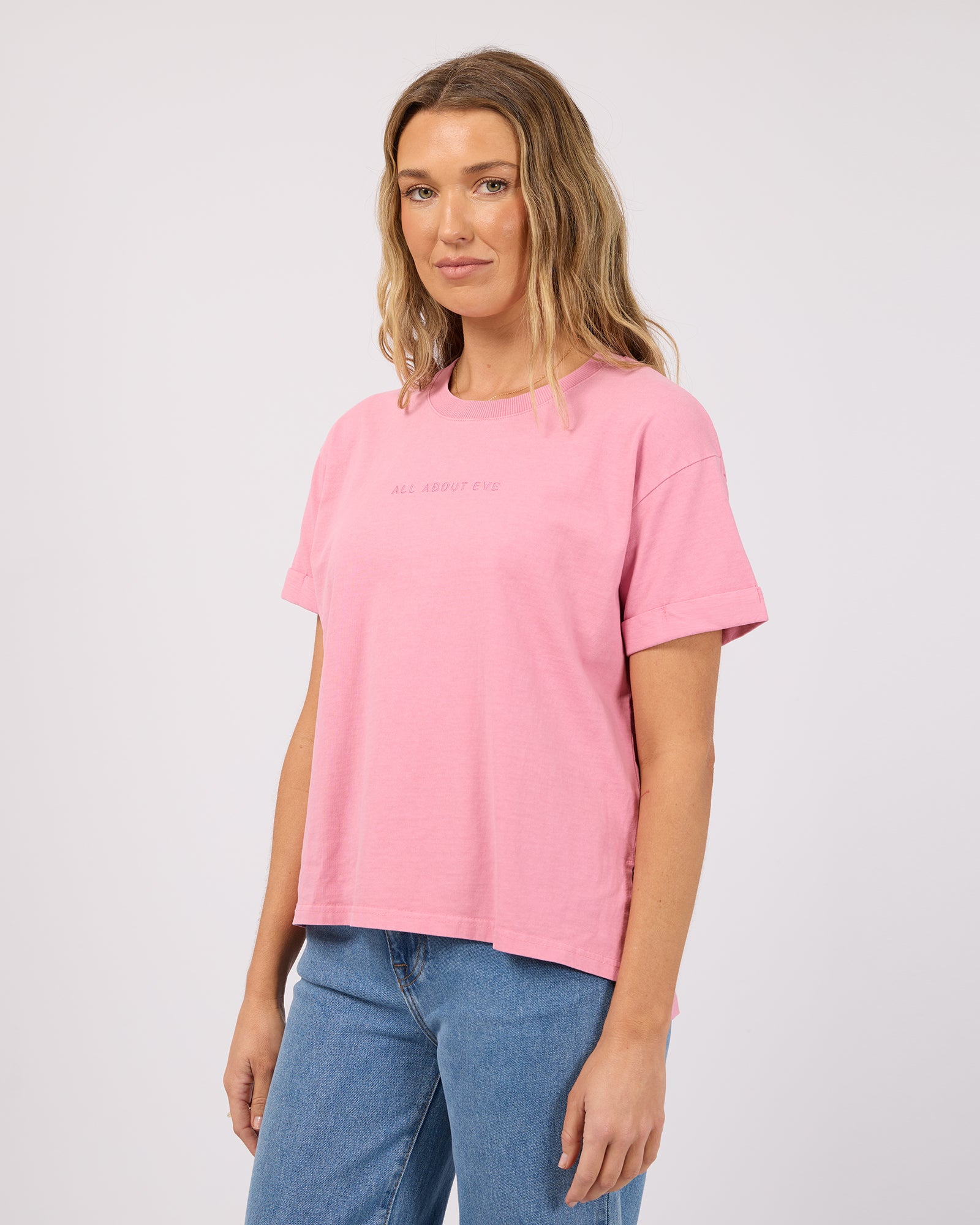 A woman modeling a light pink short-sleeve crewneck t-shirt with subtle tonal text on the chest, paired with blue jeans, against a plain light gray studio background.