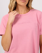 A close-up of a woman wearing a pink crewneck t-shirt with the text 'ALL ABOUT EVE' printed in matching pink on the chest, set against a plain light gray background.
