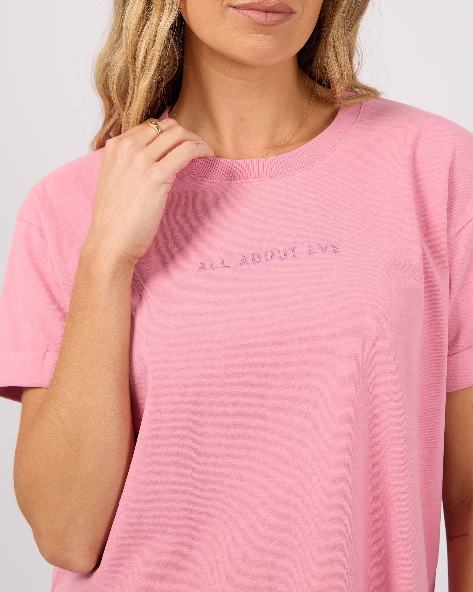 A close-up of a woman wearing a pink crewneck t-shirt with the text 'ALL ABOUT EVE' printed in matching pink on the chest, set against a plain light gray background.