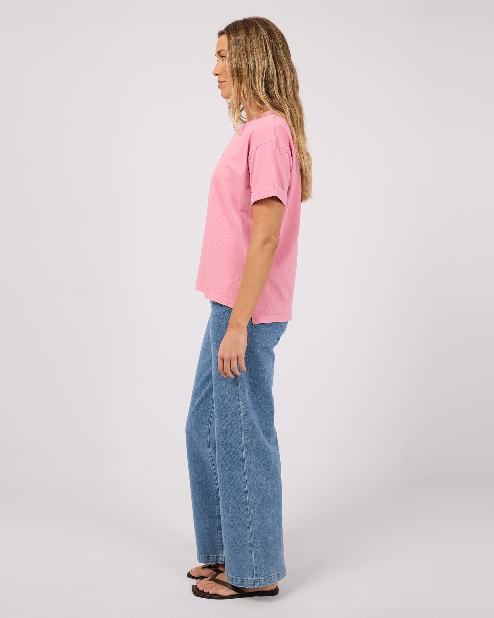 A woman shown in profile wearing a light pink short-sleeve t-shirt and wide-leg blue jeans against a plain light gray studio background.