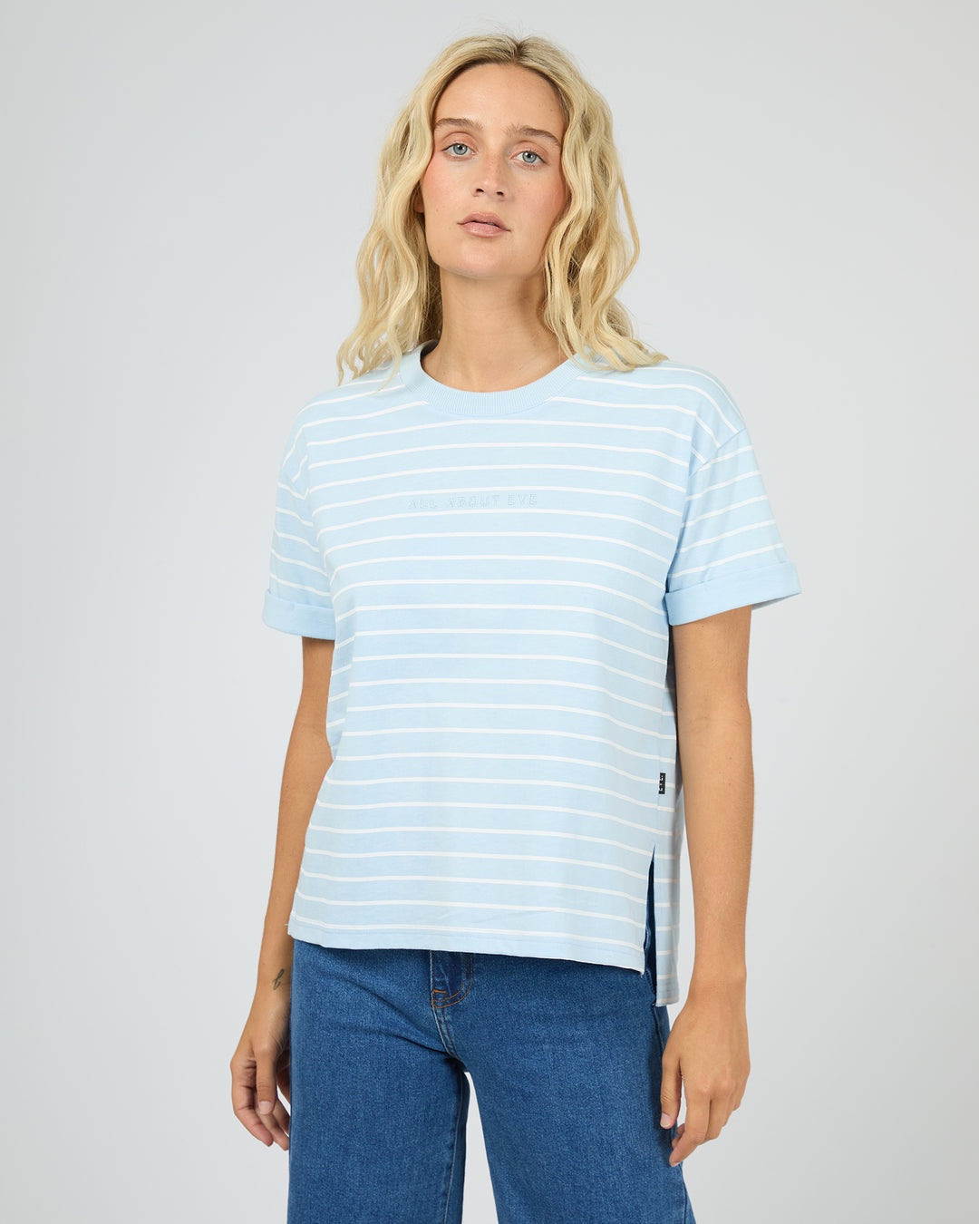 A blonde woman wearing a light blue and white horizontally striped short-sleeve t-shirt with side slits, paired with blue jeans, against a plain light gray studio background.
