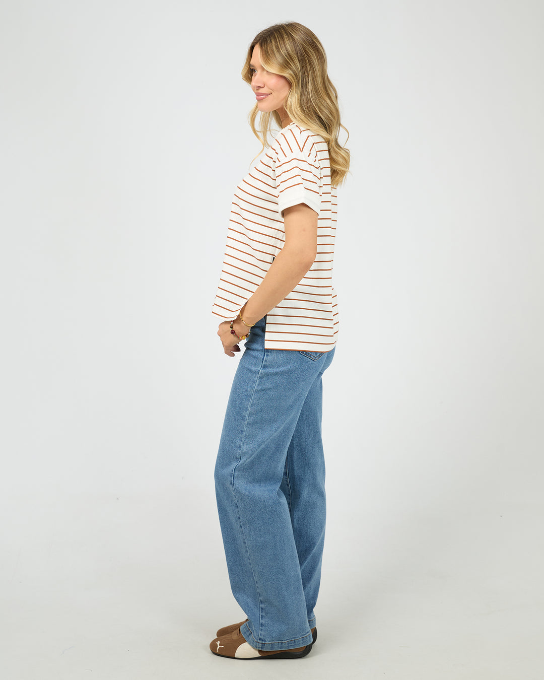 A woman modeling a white short-sleeve t-shirt with thin orange horizontal stripes, paired with wide-leg blue jeans and brown sandals, standing against a plain white studio background.