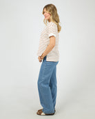 A woman modeling a white short-sleeve t-shirt with thin orange horizontal stripes, paired with wide-leg blue jeans and brown sandals, standing against a plain white studio background.