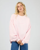 A blonde woman wearing an oversized light pink crewneck sweatshirt with subtle tonal text on the chest, paired with blue jeans, against a plain light gray studio background.