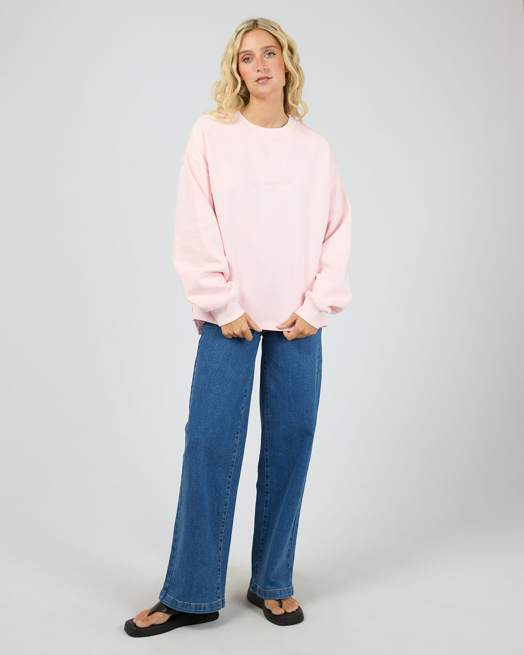 A woman modeling an oversized light pink crewneck sweatshirt with blue wide-leg jeans and black sandals, standing against a light gray studio background.