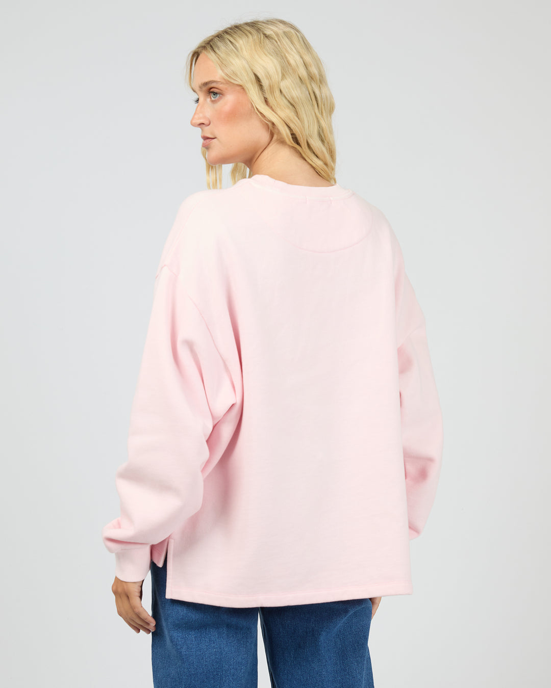 A woman with blonde hair is shown from the back wearing a light pink oversized crewneck sweatshirt and blue jeans against a light gray studio background.