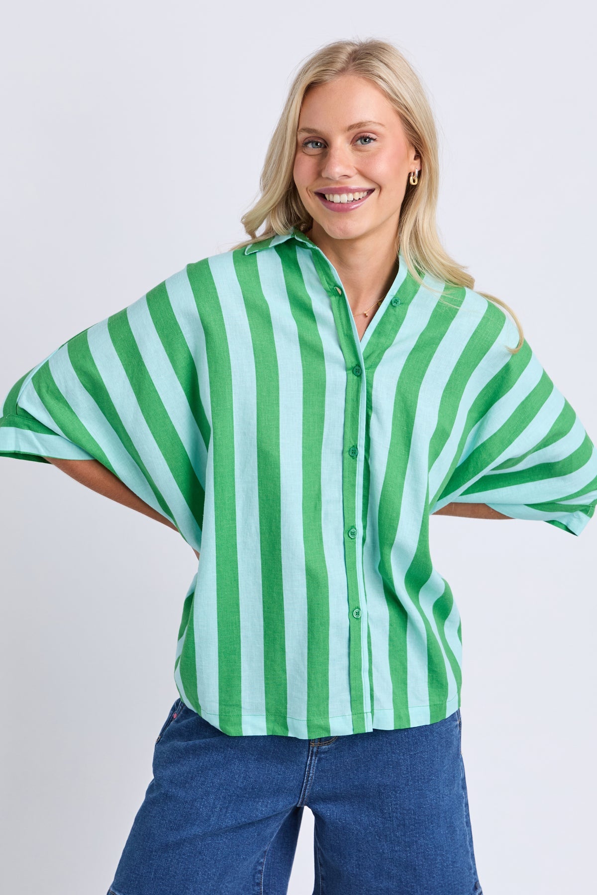 A smiling blonde woman wearing a green and white vertically striped short-sleeve button-up blouse with blue jeans, posed against a light gray studio background.