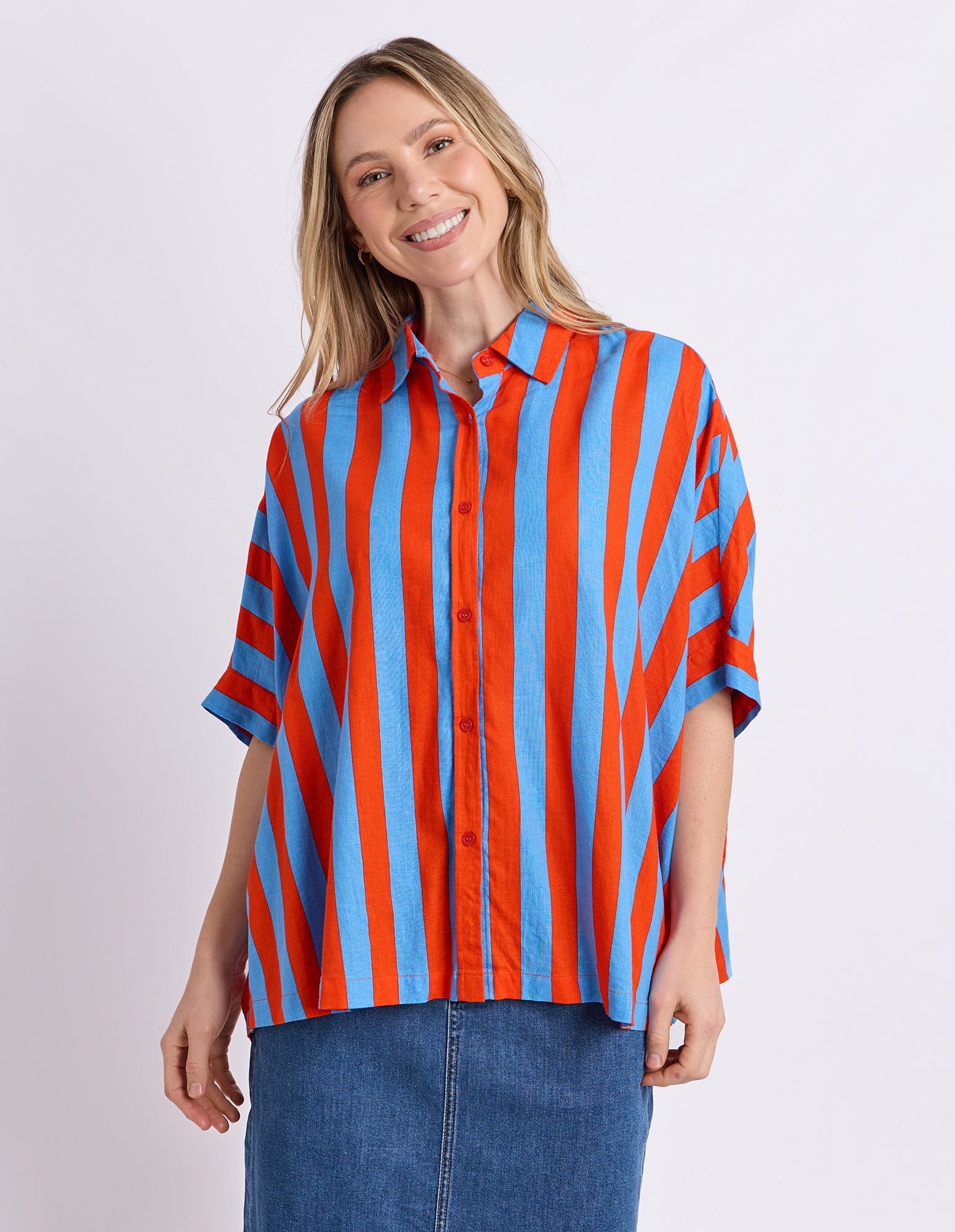 A woman modeling a short-sleeve button-up shirt with bold vertical red and light blue stripes, paired with a blue denim skirt, against a plain light gray studio background.