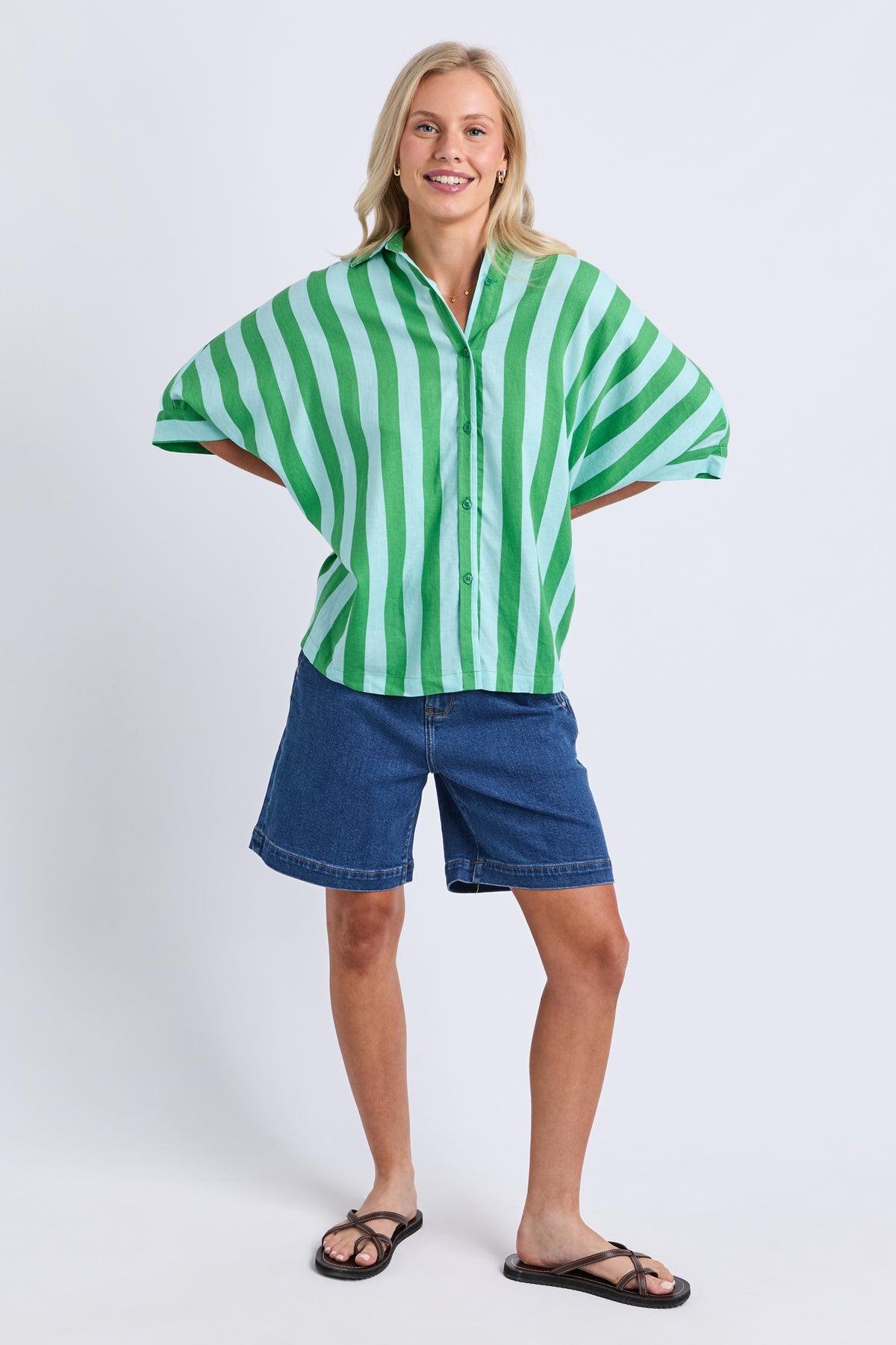 A woman modeling a green and white vertically striped short-sleeve button-up shirt with blue denim shorts and black sandals, standing against a light gray studio background.
