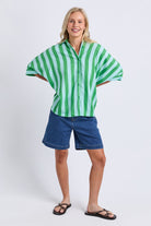 A woman modeling a green and white vertically striped short-sleeve button-up shirt with blue denim shorts and black sandals, standing against a light gray studio background.