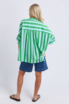 A woman shown from the back wearing a green and white vertically striped short-sleeve shirt and blue denim shorts against a light gray studio background.