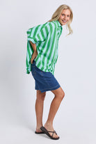 A woman modeling an oversized green and white striped button-up shirt paired with a blue denim mini skirt and black flip-flop sandals, posed against a light gray studio background.