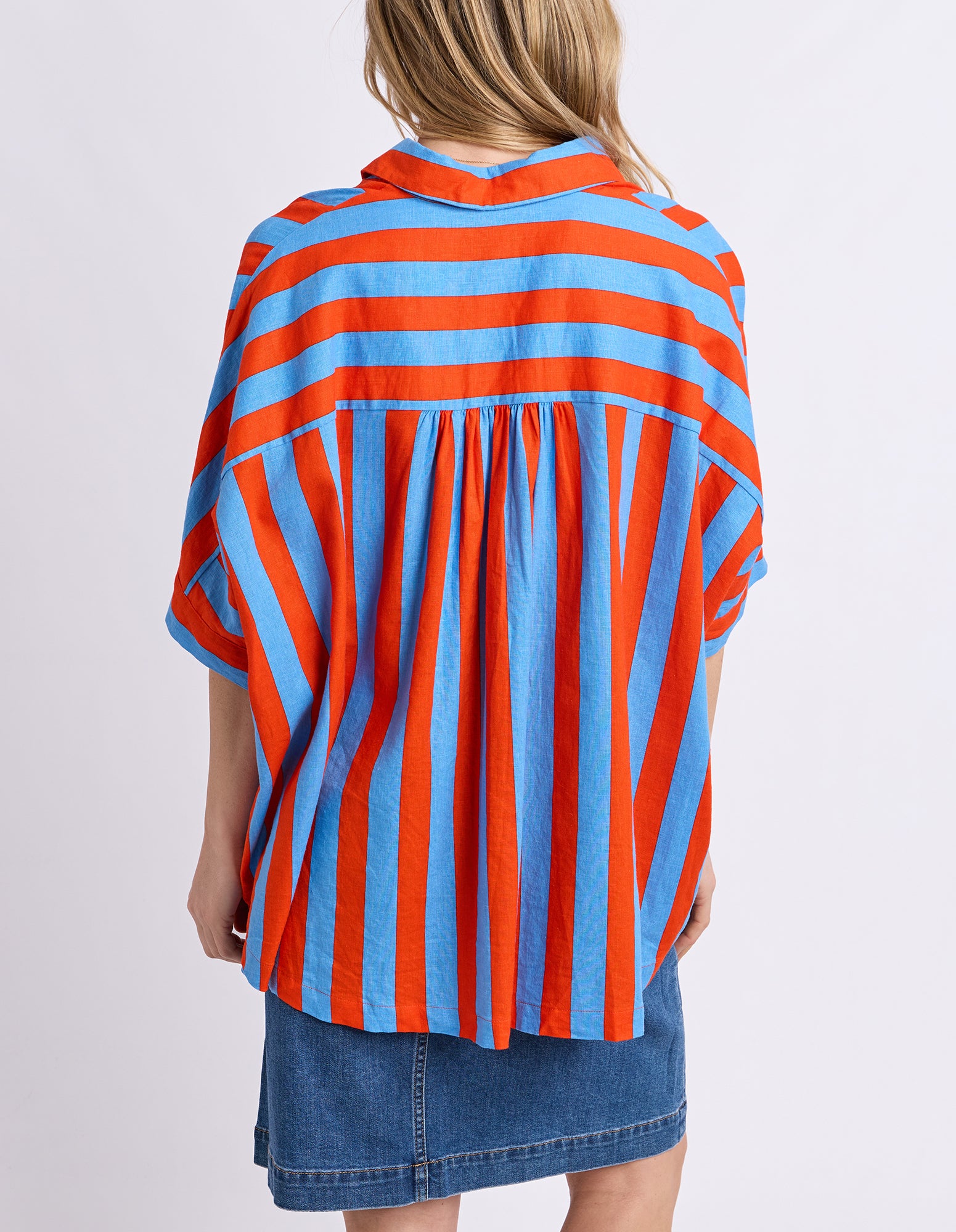 A woman shown from the back wearing a loose short-sleeve blouse with bold vertical orange and light blue stripes, paired with a denim skirt, against a plain light gray studio background.
