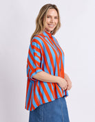 A woman wearing a vibrant blue and orange vertically striped button-up blouse with three-quarter sleeves, paired with a blue denim skirt, photographed against a light gray studio background.