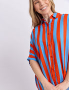 A woman wearing a short-sleeve button-up shirt with bold vertical stripes in bright orange-red and light blue, photographed against a plain light gray studio background.