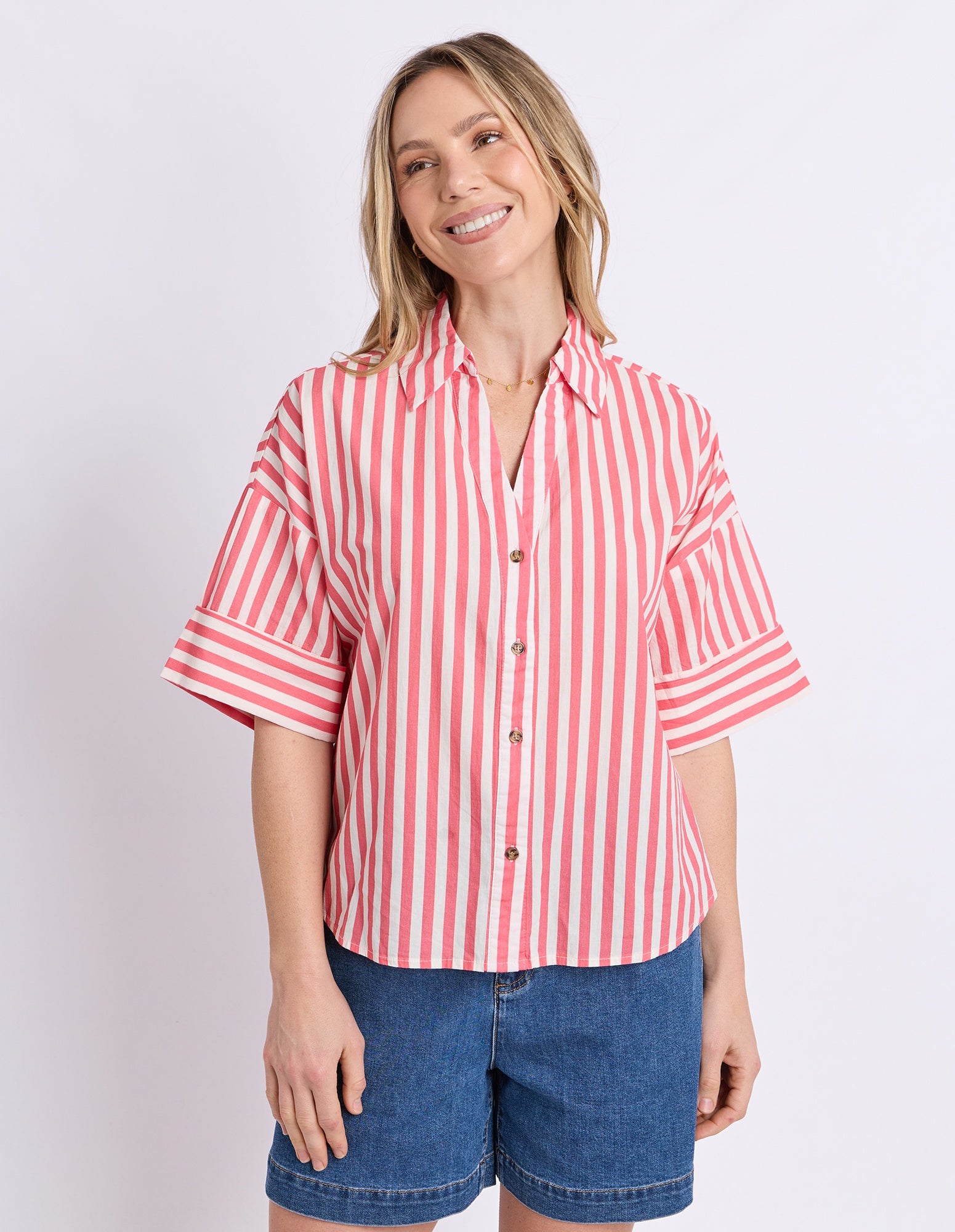 A woman modeling a short-sleeve button-up shirt with vertical pink and white stripes, paired with blue denim shorts, against a light gray studio background.