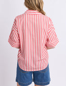 A woman viewed from the back wearing a short-sleeve collared button-up shirt with vertical pink and white stripes, paired with blue denim shorts, against a plain light gray studio background.