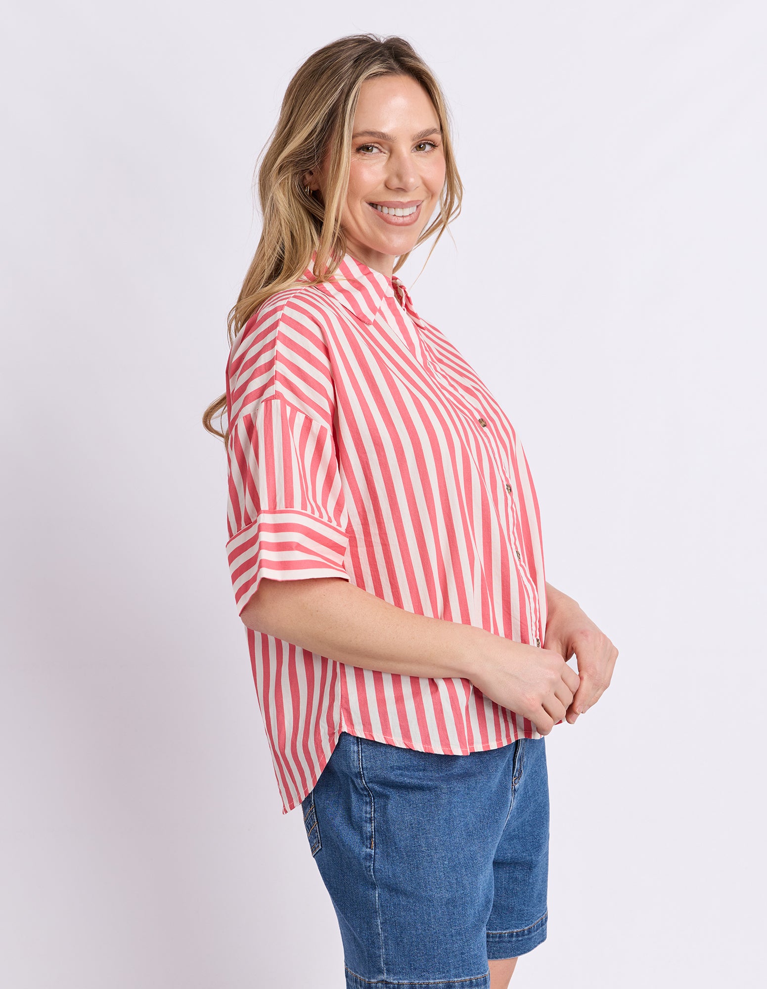 A woman modeling a short-sleeve button-up shirt with vertical red and white stripes, paired with blue denim shorts, against a plain light gray background.