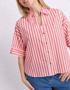 A woman wearing a pink and white vertically striped short-sleeve button-up shirt with rolled cuffs, paired with blue jeans, against a light gray studio background.