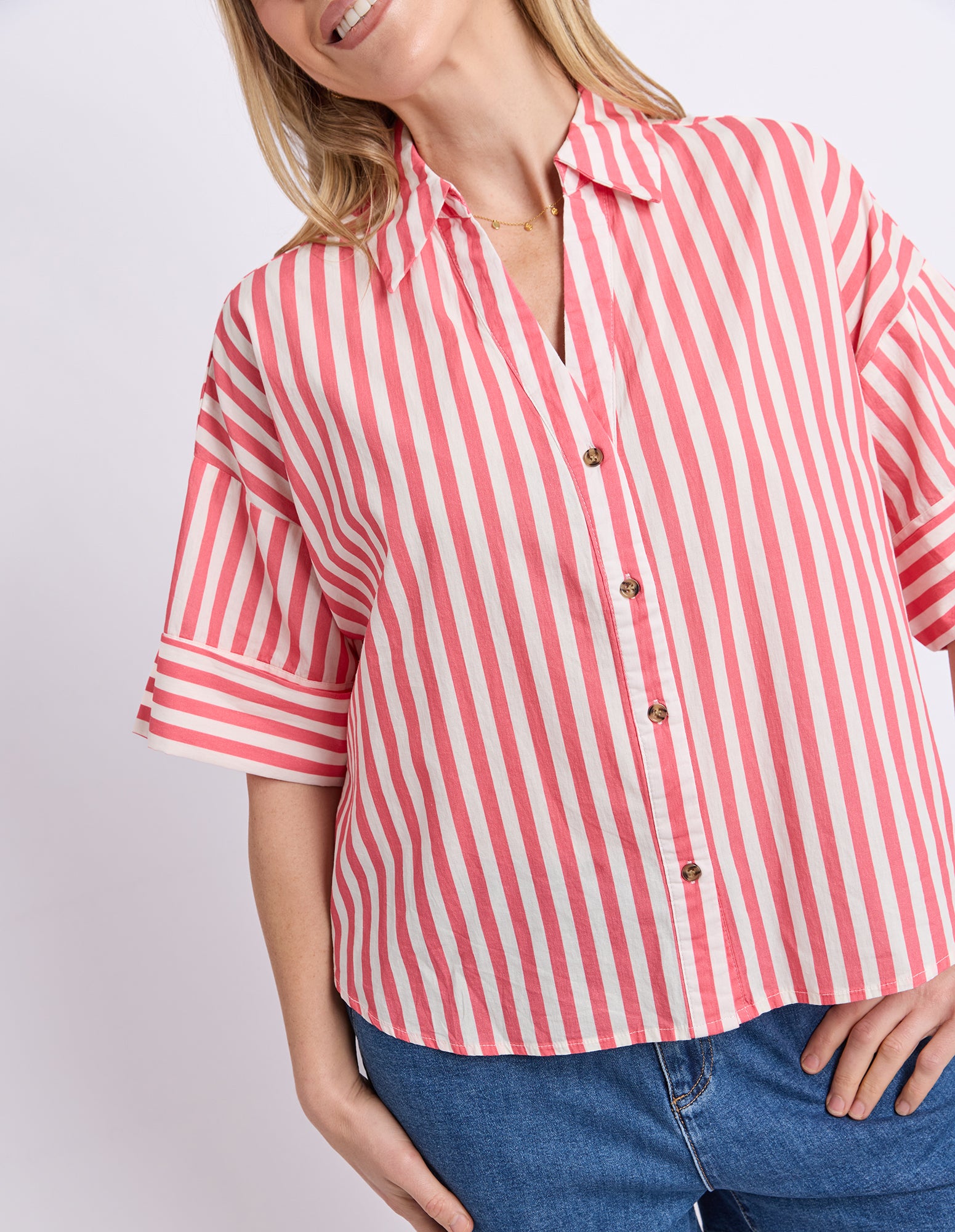 A woman wearing a pink and white vertically striped short-sleeve button-up shirt with rolled cuffs, paired with blue jeans, against a light gray studio background.