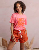 Woman wearing a pink t-shirt with orange graphics and red shorts against a beige background