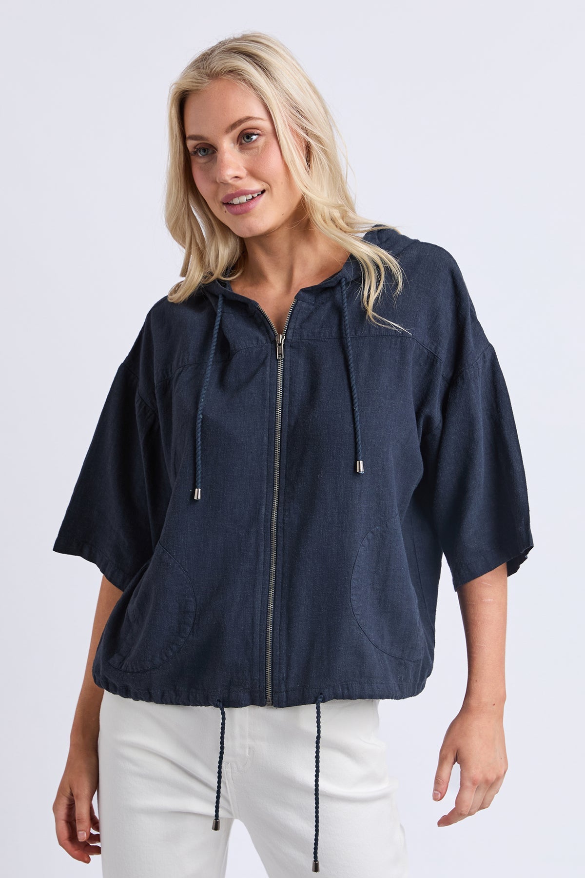 A woman wearing a navy blue zip-up hooded jacket with three-quarter sleeves and white pants, photographed against a light gray studio background.