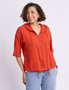 A woman wearing a bright orange hooded zip-up jacket with three-quarter sleeves and drawstrings, paired with light blue jeans, photographed against a light gray studio background.
