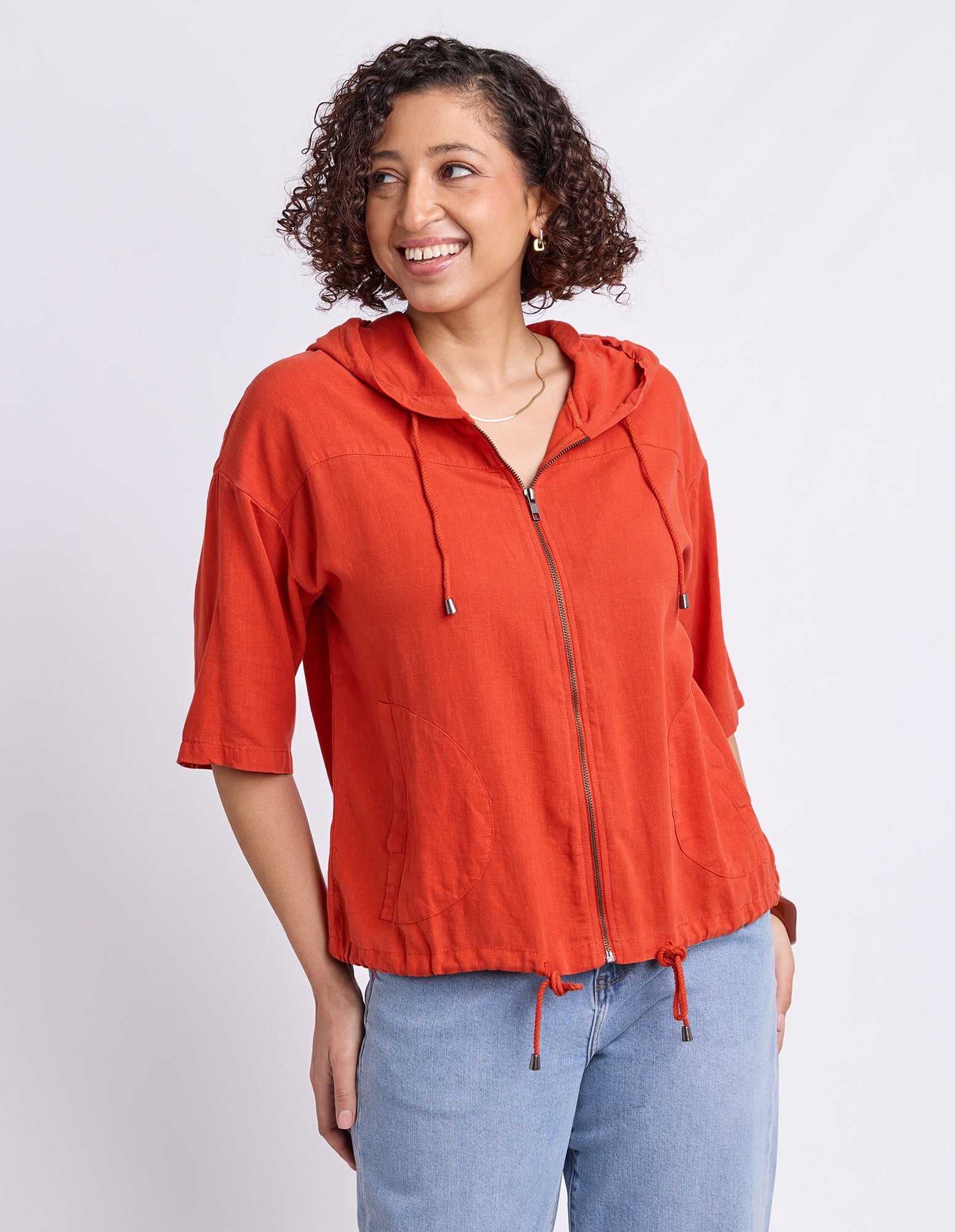 A woman wearing a bright orange hooded zip-up jacket with three-quarter sleeves and drawstrings, paired with light blue jeans, photographed against a light gray studio background.