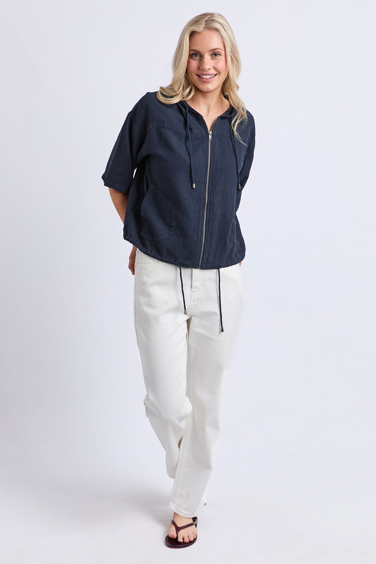 A woman modeling a navy blue short-sleeve zip-up hoodie with white drawstring pants and dark sandals, standing against a light gray studio background.