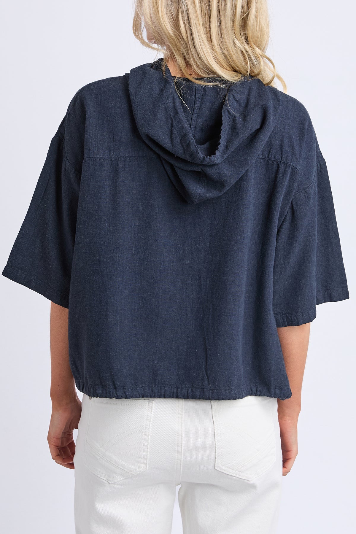 A woman viewed from the back wearing a navy blue cropped short-sleeve top with a draped hood and white pants, against a plain light gray studio background.