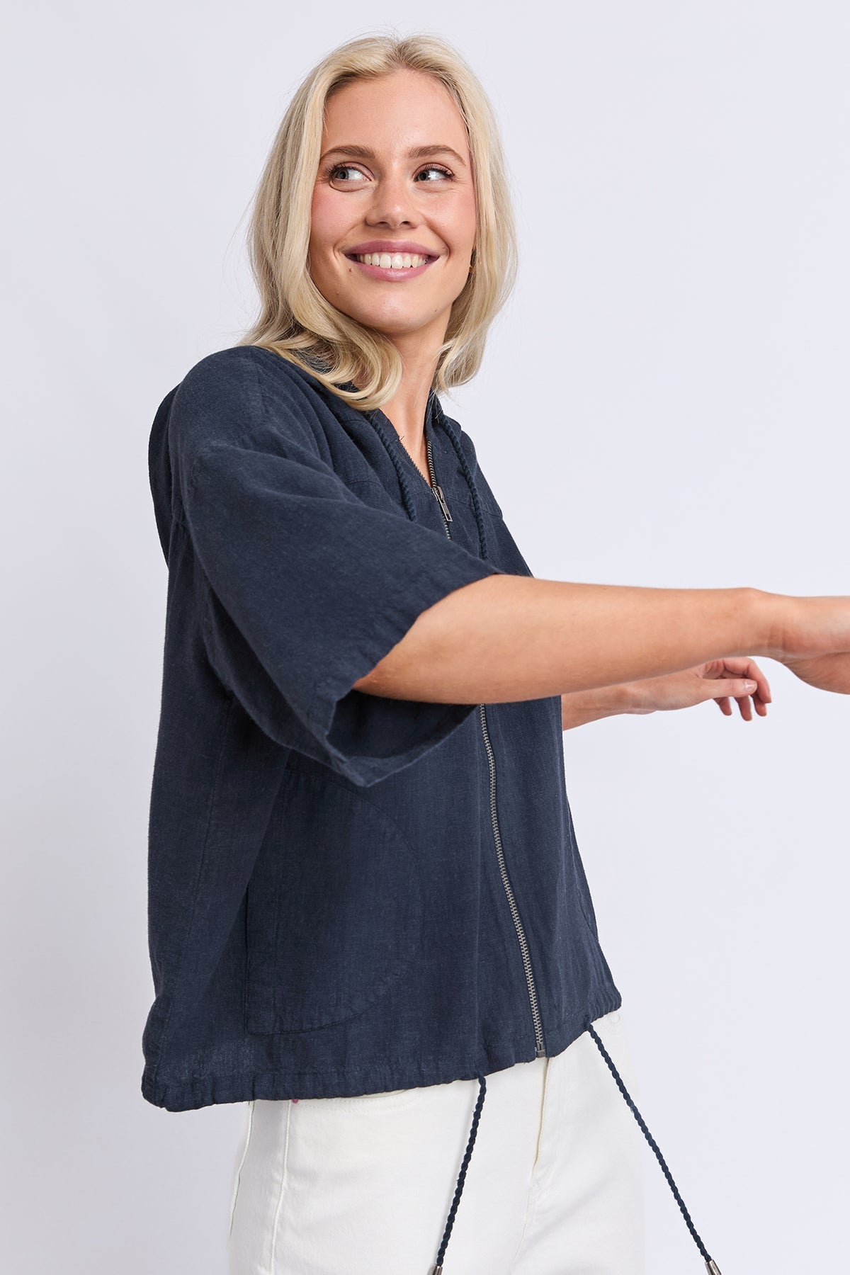 A smiling blonde woman wearing a dark navy blue linen-blend zip-up jacket with white pants, shown from the waist up against a light gray studio background.