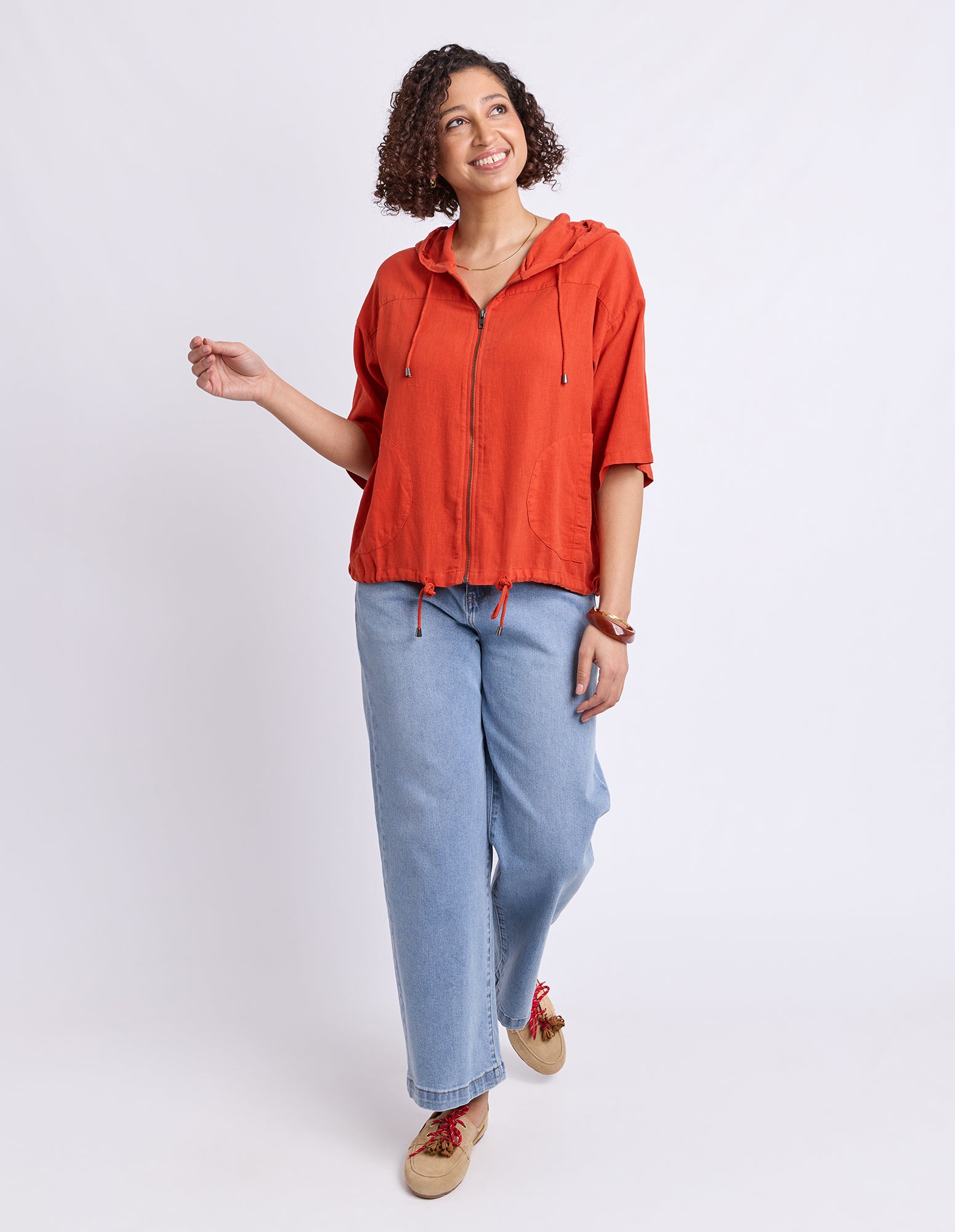 A woman modeling a rust-orange zip-up hooded sweatshirt with light-wash blue jeans and tan sneakers, standing against a plain light gray studio background.