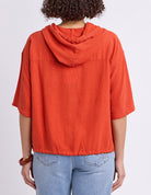 A person shown from the back wearing a bright orange hooded short-sleeve top and light blue jeans against a plain light gray studio background.