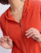 Close-up of a person wearing a bright orange zip-up hooded jacket, holding the drawstrings near their hands against a plain light background.