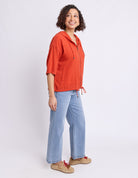 A woman modeling a bright orange hooded zip-up jacket with light blue jeans and tan shoes, standing against a plain white studio background.