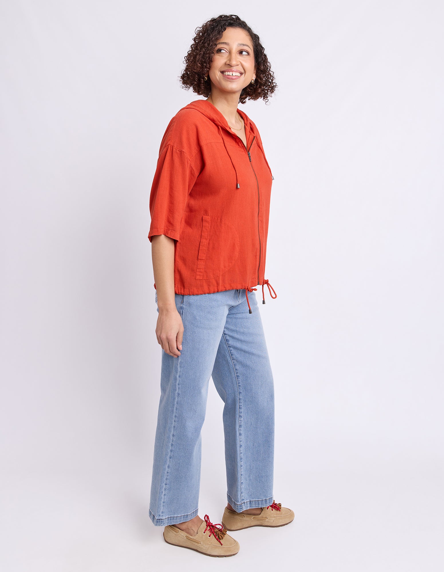 A woman modeling a bright orange hooded zip-up jacket with light blue jeans and tan shoes, standing against a plain white studio background.