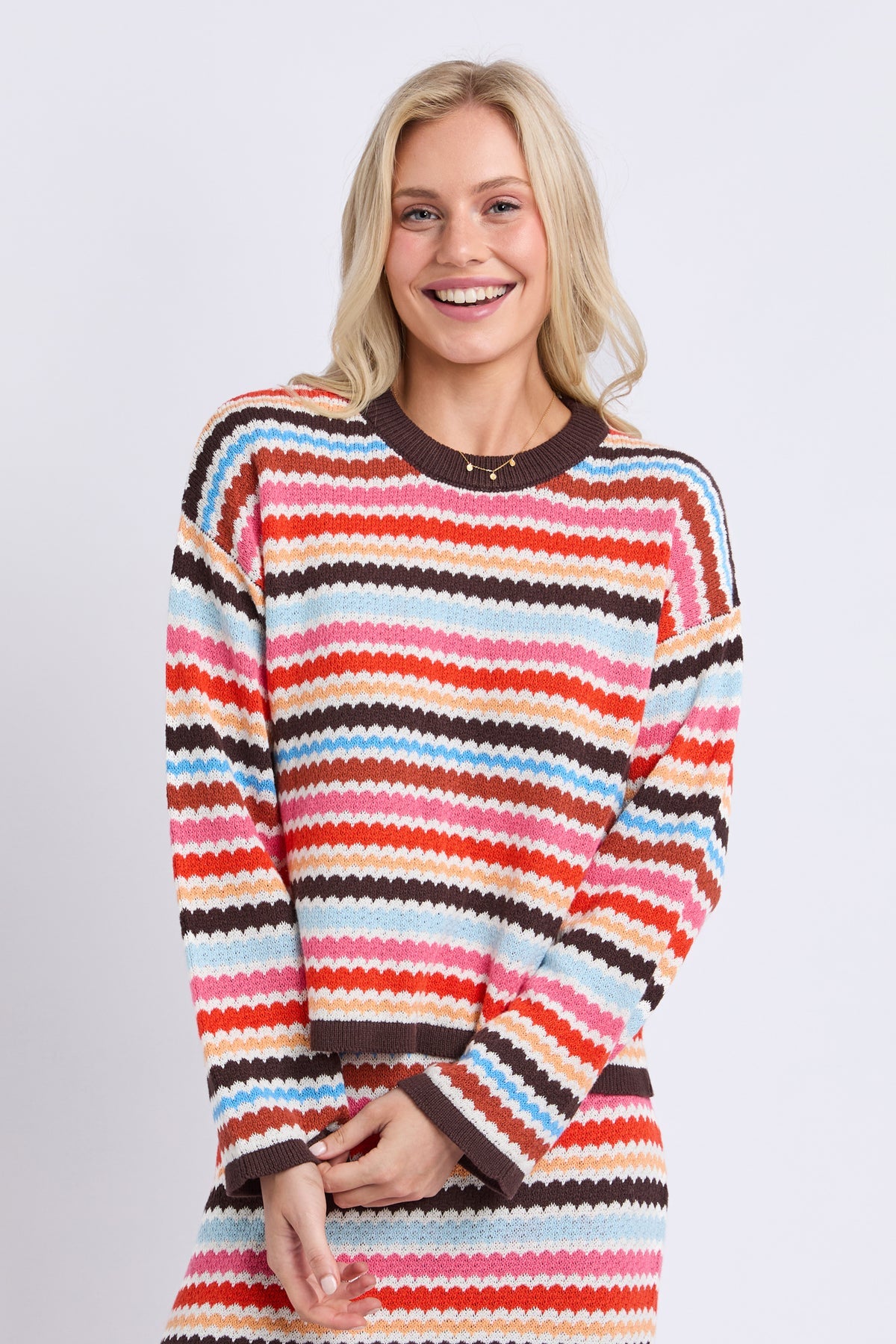 A smiling blonde woman wearing a long-sleeve crewneck sweater with horizontal wavy stripes in orange, red, pink, light blue, cream, and dark brown, photographed against a light gray studio background.