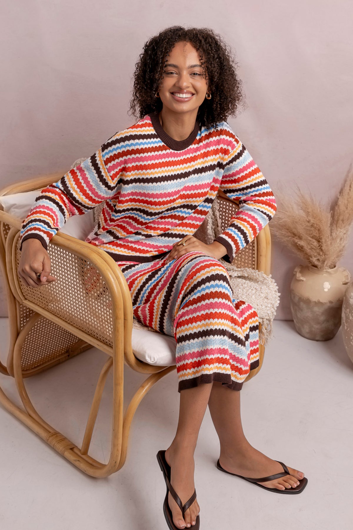 A woman wearing a matching long-sleeve knit top and cropped pants set with horizontal multicolor stripes in red, orange, yellow, white, and dark brown, sitting in a rattan chair against a soft beige background.