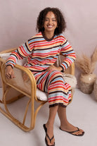 A woman wearing a matching long-sleeve knit top and cropped pants set with horizontal multicolor stripes in red, orange, yellow, white, and dark brown, sitting in a rattan chair against a soft beige background.