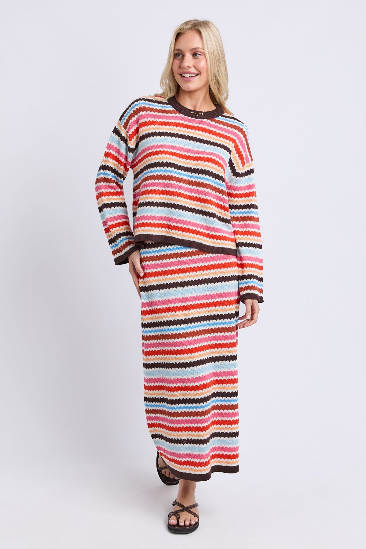 A woman modeling a matching knit two-piece set featuring a cropped long-sleeve top and a maxi skirt, both decorated with horizontal wavy stripes in red, orange, pink, light blue, black, and white, set against a plain light gray background.