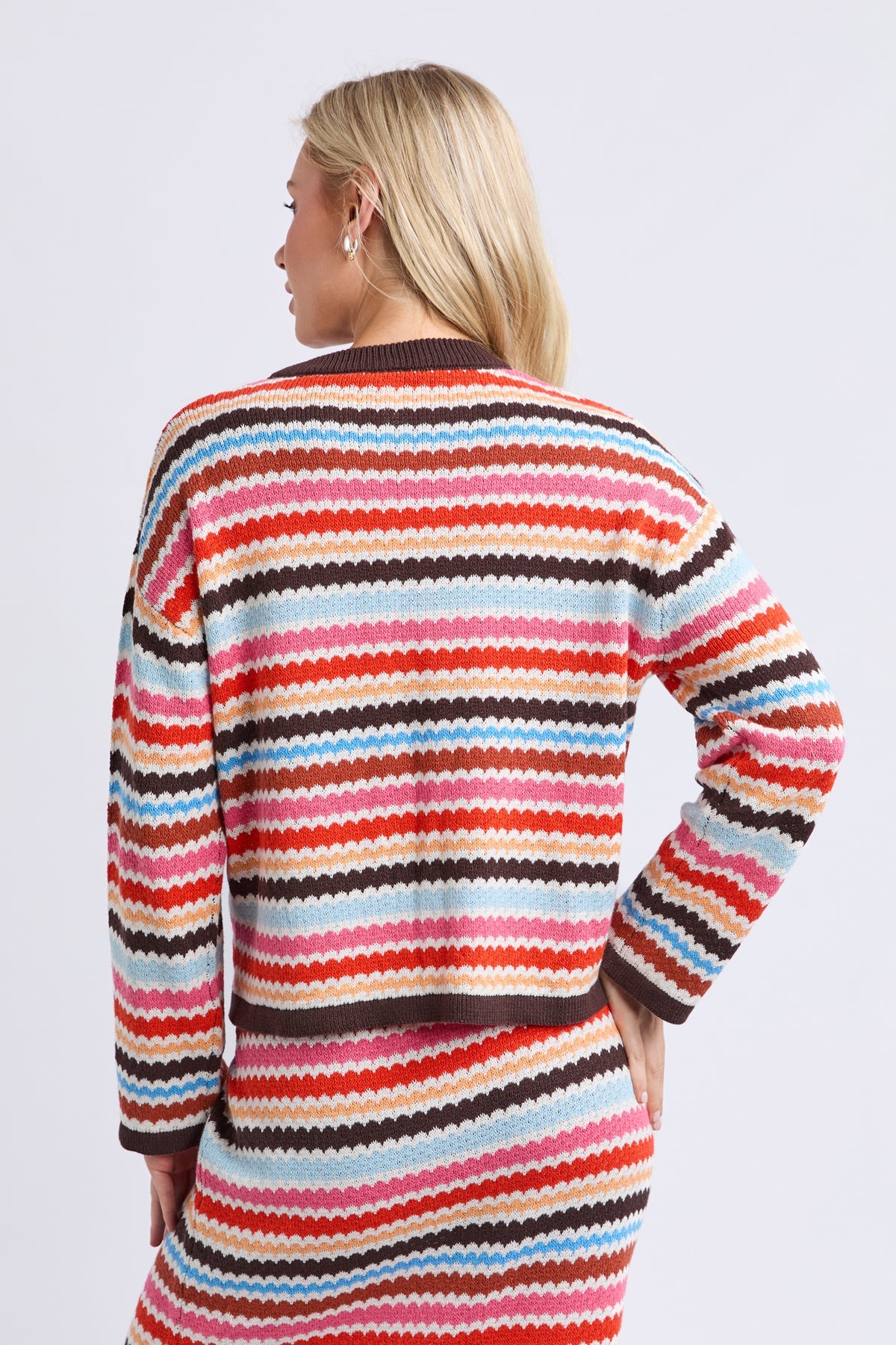 A woman shown from the back wearing a long-sleeve knit sweater dress with horizontal wavy stripes in red, orange, pink, cream, light blue, and dark brown on a light gray studio background.