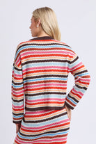 A woman shown from the back wearing a long-sleeve knit sweater dress with horizontal wavy stripes in red, orange, pink, cream, light blue, and dark brown on a light gray studio background.