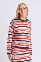 A smiling blonde woman wearing a colorful striped knit sweater and matching striped skirt against a light gray studio background.