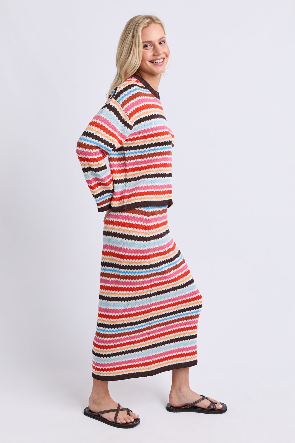 A woman modeling a long-sleeve knit maxi dress with horizontal wavy stripes in red, orange, pink, black, and light blue, shown from a side-back angle against a light gray studio background.